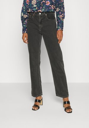 Frau trägt schwarze, gerade Jeans mit hoher Taille, ein langärmeliges Oberteil mit Blumenmuster und schwarze Sandalen mit offenem Zehenbereich und Absatz.