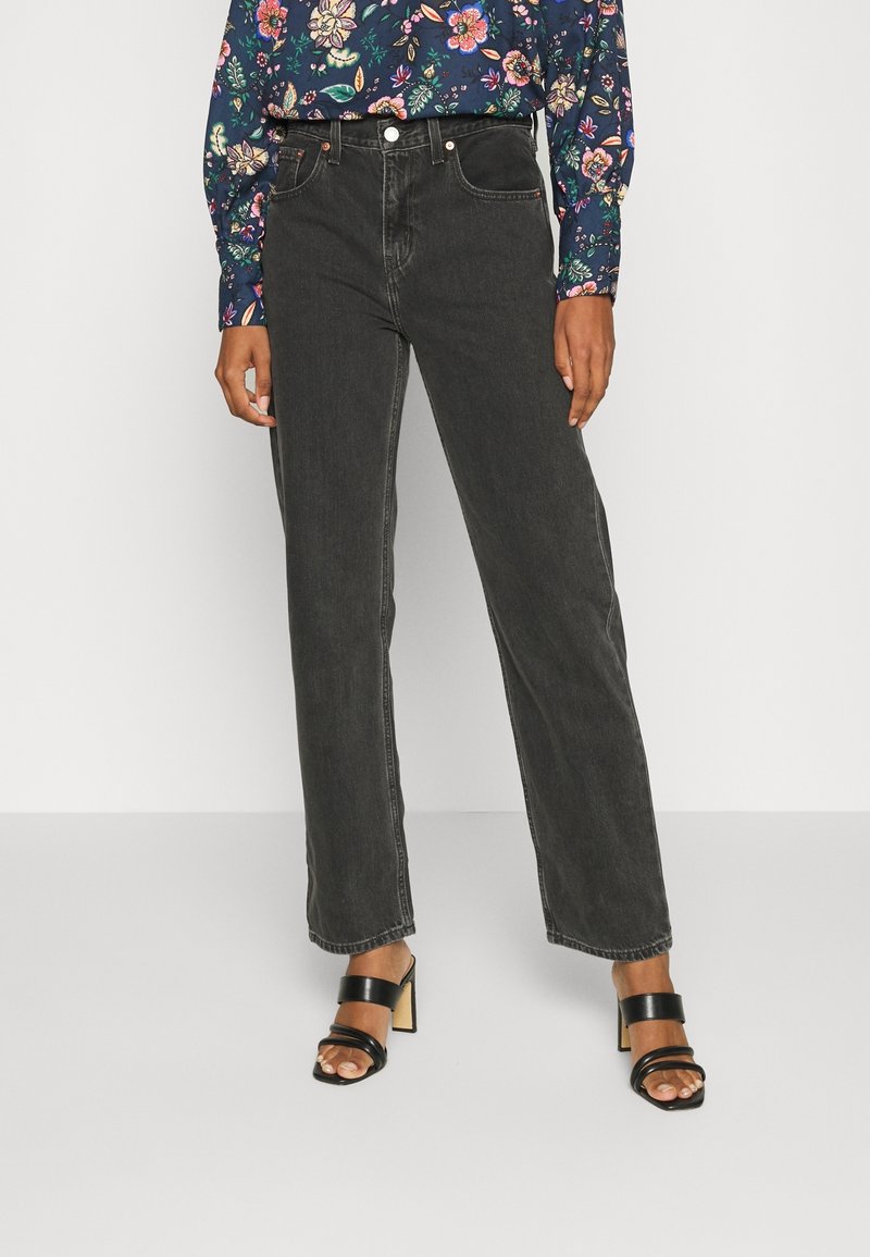 Femme portant un jean droit noir taille haute, un haut fleuri à manches longues et des sandales à talons noires ouvertes à l’avant.