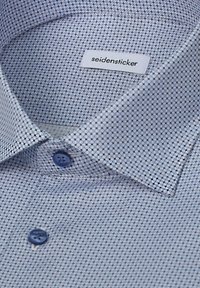 Gros plan d'une chemise habillée bleu clair à motif pliée, avec des boutons bleus et une étiquette de marque "seidensticker" visible à l'intérieur du col.