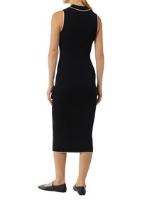 Schwarzes, geripptes Midikleid mit taillierter Form, ärmellosem Design und weißen Paspelierungen am Ausschnitt und Saum. Schlichte flache schwarze Schuhe.