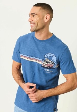 Lächelnder Mann im blauen T-Shirt mit Vintage Volkswagen-Van-Grafik, der durch eine Welle und pastellfarbene Streifen fährt.