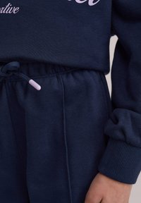 Tums zila sporta kostīma komplekts ar mīkstu tekstūru. Ir ribota aproce un jostasvietā aukla ar gaiši rozā aproci. Uzraksts gaiši rozā krāsā.