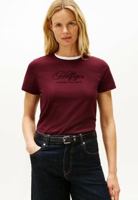 Bordeaux kortærmet t-shirt med hvid halsudskæring og sort logotekst. Kombineret med mørke denim jeans og et sort bælte.