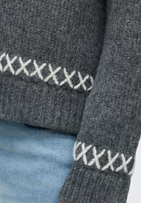 Grauer gestrickter Pulloverärmel und Bund mit weißem Kreuzstich-Detail, kombiniert mit hellblauen Jeans aus Denim.