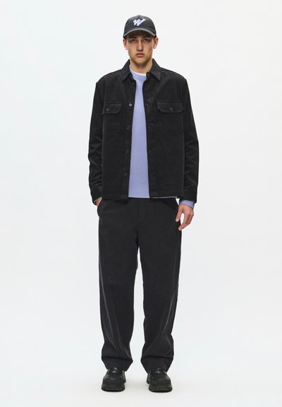 Jeune homme debout, portant une casquette noire, une veste noire boutonnée, un pull bleu clair, un pantalon noir ample et des chaussures noires sur fond blanc.