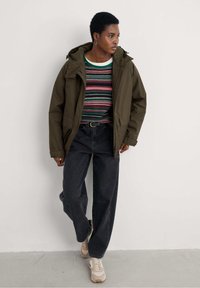 Oliwkowa wodoodporna kurtka z kapturem, zestawiona z wielokolorową, w paski długą bluzką, czarnymi, luźnymi dżinsami i beżowymi sneakersami.