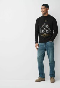 Čierny sveter s vianočným dizajnom pív v pohároch usporiadaných ako vianočný stromček, s farebnými akcentmi a textom "Je čas na Guinness".