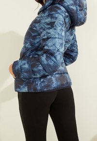 Puffjakke i blå tie-dye mønster med hætte, quiltet tekstur og elastiske manchetter. Sammen med sorte leggings.