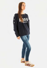Marineblauer Pullover mit dem Schriftzug "PROUD", Knopfdetails an den Ärmeln, kombiniert mit blauen Jeans und beigen Sneakers mit bordeauxroten Streifen.