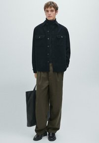 Giacca camicia in velluto nero con bottoni a pressione, due tasche sul petto, abbinata a pantaloni larghi verde oliva e scarpe in pelle nere. Borsa tote nera.