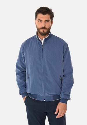 Uomo con la barba che indossa una giacca bomber blu con la cerniera chiusa e due tasche frontali, in piedi davanti a uno sfondo chiaro e uniforme.