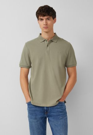 Junger Mann steht mit den Händen in den Taschen, trägt ein olivgrünes Poloshirt und blaue Jeans vor einem schlichten hellgrauen Hintergrund.