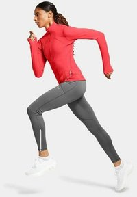Rode zip-up sporttop en grijze legging, met een aansluitend ontwerp. Model in hardloophouding, draagt witte sneakers met reflecterende accenten.