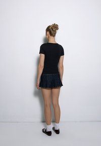 Czarna dopasowana koszulka zestawiona z ciemnoniebieską mini spódnicą z denimu. Modelka nosi białe skarpetki do kostek i brązowe buty, widoczna z tyłu na tle gładkiej ściany.