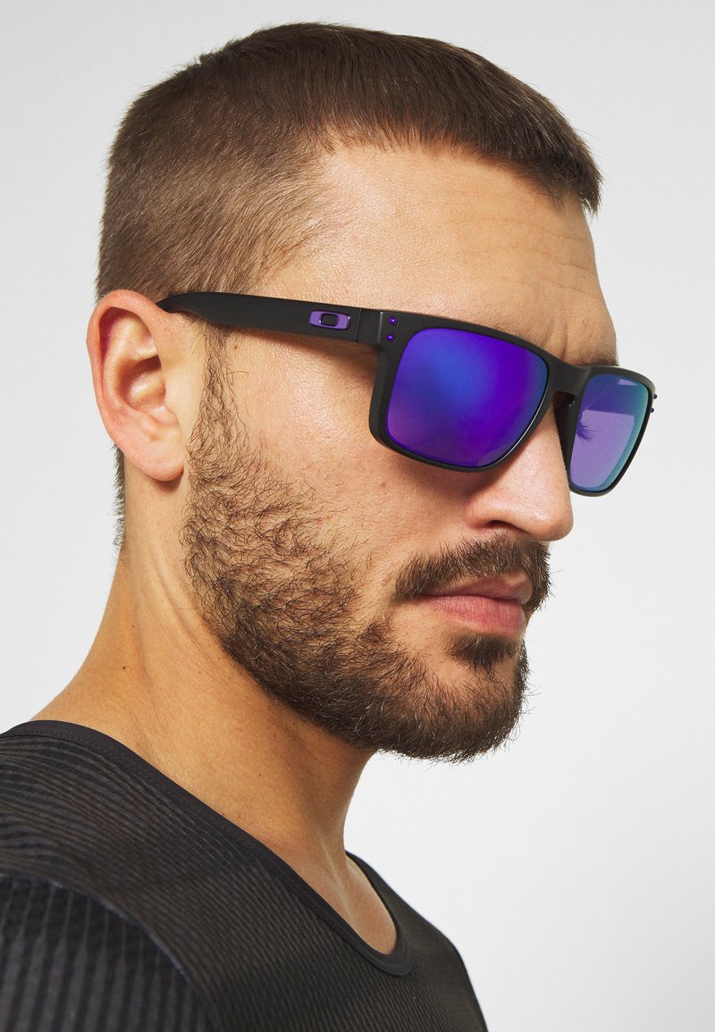 Schwarze Sonnenbrille mit lila verspiegelt Gläsern, quadratischem Rahmen, strukturierten Bügeln und einem kleinen Logo-Detail an der Seite.