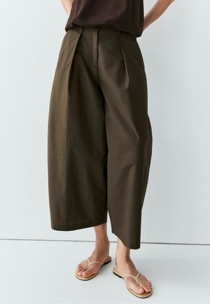 Person trägt lockere, braune, weite, knöchellange Hosen und beige flache Zehensandalen und steht vor einem schlichten weißen Hintergrund.
