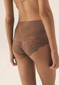 Kobieta w brązowej, wysokiej bieliźnie koronkowej, stojąca z rękami luźno wzdłuż ciała, widocznym tatuażem serca na lewym ramieniu.