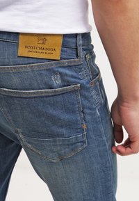 Vaqueros de mezclilla azul con un parche de cuero con la etiqueta "Scotch & Soda", que presentan un desgaste sutil, detalles de costura amarilla y un corte ajustado.