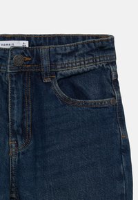 Blaue Denim-Jeans mit dunkler Wäsche, ausgestattet mit gelben Nähten, einem Knopfverschluss und Fronttaschen. Das Material ist weich und strukturiert.