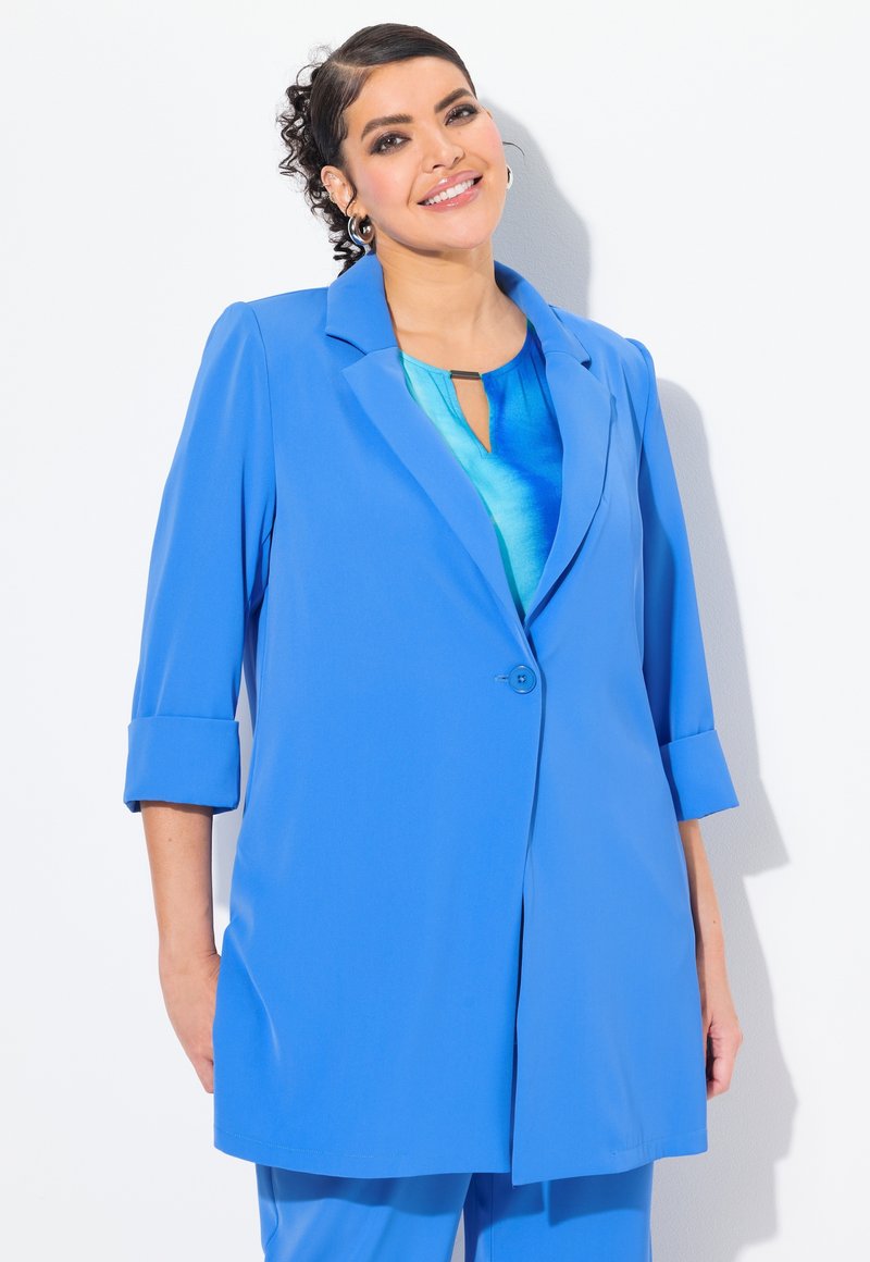 Vaaleansininen bleiseri, joka on valmistettu sileästä kankaasta, jossa on yksittäinen napin sulku, lovettu kaulus ja käärityt hihat, yhdistetty siniseen paitaan.