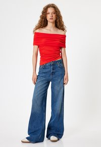 Rotes, schulterfreies, gerafftes Oberteil kombiniert mit weiten, verblassten blauen Denim-Jeans. Braune Sneaker betonen das Outfit. Einfaches, lässiges Design.