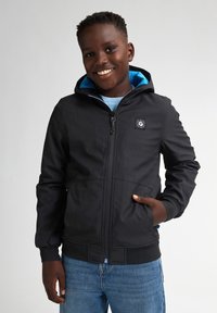 Schwarze Zip-Jacke mit Kapuze, ausgestattet mit blauen Innenfutter, elastischen Bündchen und einem Logopatch auf der Brust. Getragen über einem blauen Oberteil und Jeans.
