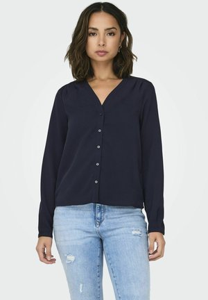 Blouse bleu marine à manches longues avec un col en V et une fermeture à boutons, fabriquée dans un tissu lisse. Portée avec un jean bleu déchiré, affichant un look décontracté.