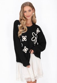 Czarny sweter z dzianiny z białymi, kwiatowymi wzorami, w oversize'owym fasonie, z teksturowanej tkaniny, połączony z białą sukienką z koronkowymi wykończeniami. Długie rękawy widoczne.
