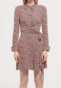 Robe à motifs rouges et blancs avec col, manches longues et ceinture à nouer à la taille. Présente un volant à l'avant et une jupe évasée.