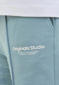 Svijetloplave trenirke od mekog materijala s bočnim džepovima. Sadrže bijeli tekst: "Originals Studio" i detalje o marki u jednostavnom fontu.