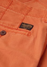 Pomarańczowa tkanina o gładkiej fakturze, z czarną metką z napisem "Superdry" i guzikami. Zawiera przeszycia i detal w postaci kieszonki naszywanej.