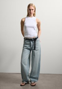 Weißes ärmelloses Oberteil, hellblaue, weit geschnittene Jeans mit einem schwarzen Gürtel und einer Lasche. Die Jeans haben eine ausgewaschene Textur und einen lässigen Sitz.