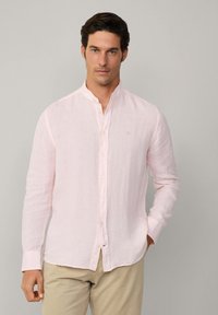 Camisa de lino color rosa claro con cuello mandarín, un bolsillo y botones blancos. Mangas largas con puños enrollados. Diseño minimalista.