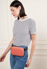 Borsa a tracolla corallo con design strutturato, realizzata in materiale testurizzato, chiusura con zip, attaccata a una cintura nera sopra jeans blu e una maglietta a righe.