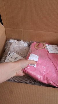 Roupa rosa com uma alça arredondada, parte de uma parte superior riscada visível sob uma embalagem transparente, dentro de uma caixa de papelão marrom.