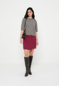 Γκρι ζακέτα με κοντά μανίκια και κουμπιά, συνδυασμένη με μπορντό φούστα τύπου wrap και μαύρες μπότες έως το γόνατο. Απλός σχεδιασμός με ελάχιστες λεπτομέρειες.