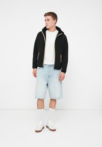 Giovane uomo che indossa una giacca nera con zip sopra una maglietta bianca, pantaloni denim leggeri blu chiaro, calzini bianchi e sneakers bianche, in piedi su uno sfondo bianco.