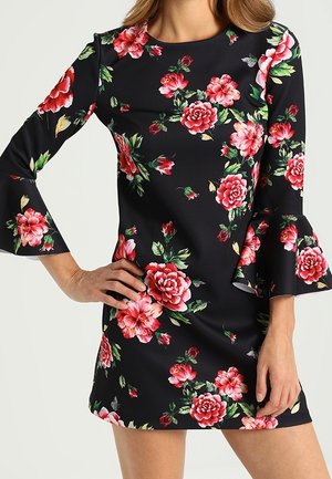 Vrouw in een zwarte jurk met een rood en roze bloemenpatroon, met lange klokmouwen en een getailleerd silhouet.