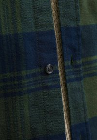 Nahaufnahme eines grün- und marineblauen karierenden Stoffes mit einem dunklen Knopf und einem beigen Strickbesatz am Rand.