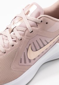 Baskets de sport rose pâle en matière en maille, avec des accents texturés, un logo central en forme de swoosh en or métallique et une semelle blanche contrastante.