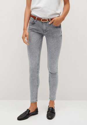 Personne portant un jean skinny gris clair avec une ceinture marron, un haut blanc, et des chaussures noires sans lacets, debout devant un fond uni.