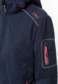 La chaqueta softshell en color navy cuenta con un bolsillo con cremallera en el brazo con acentos en rosa, ribetes grises y un logo en el hombro; tiene tejido texturizado y capucha.