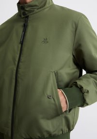 Olive green bomber jacket with a stand collar, front zipper, and side pockets. Features a subtle logo and ribbed cuffs. Smooth, glossy fabric.