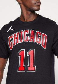 Camiseta de algodón negra con "CHICAGO" en rojo, con el número "11" debajo y un logo de Jordan en blanco en el hombro izquierdo.
