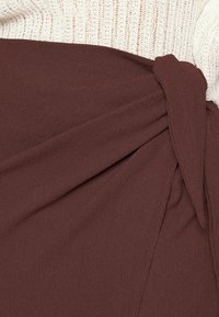 Bruine stoffen gordijnen met een knoopdetail op de taille, aangevuld met een gemêleerde crèmekleurige bovenkant, wat een gelaagd uiterlijk creëert.