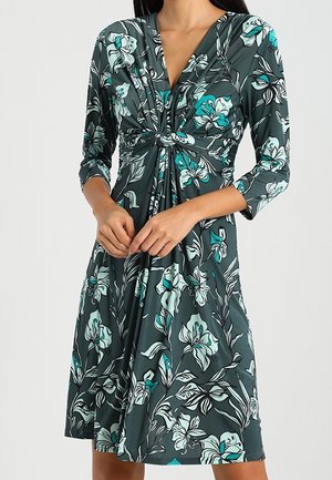 Femme portant une robe florale vert foncé longueur genou avec manches trois-quarts et un détail de nœud torsadé au niveau de la poitrine.