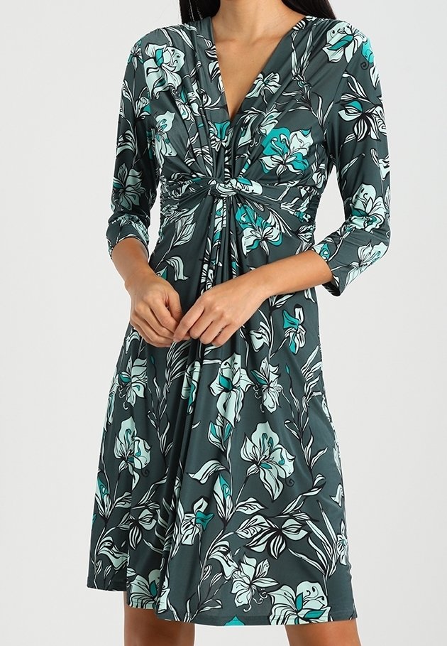 Femme portant une robe florale vert foncé longueur genou avec manches trois-quarts et un détail de nœud torsadé au niveau de la poitrine.