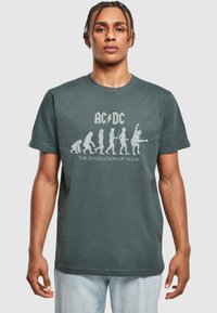 Mørkegrøn bomulds t-shirt med hvidt AC/DC grafik kunstværk, der skildrer udviklingen af rockdesign. Korte ærmer, rund halsudskæring, afslappet pasform.