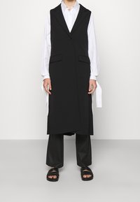 Zwart mouwloos vest met een gestructureerd ontwerp, voorzien van twee voorzakken en een enkele knoopsluiting, gecombineerd met losse zwarte broek en sandalen.