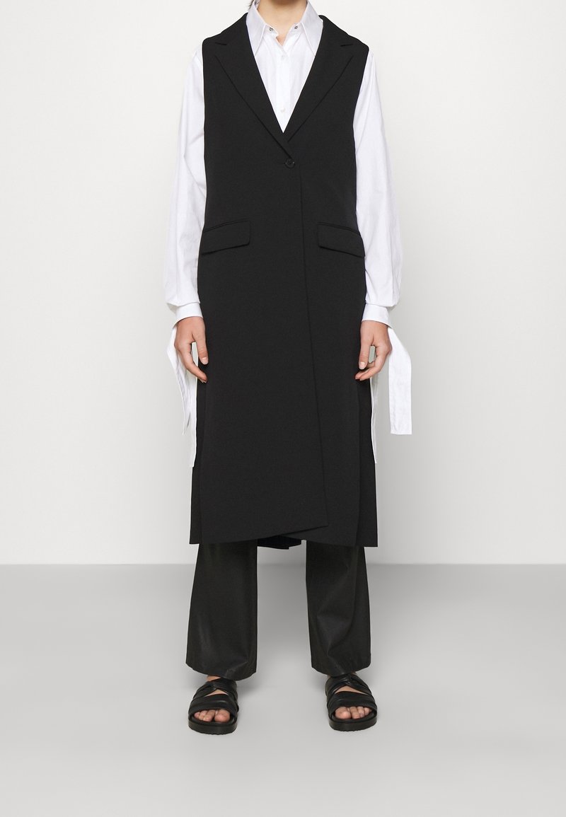 Zwart mouwloos vest met een gestructureerd ontwerp, voorzien van twee voorzakken en een enkele knoopsluiting, gecombineerd met losse zwarte broek en sandalen.